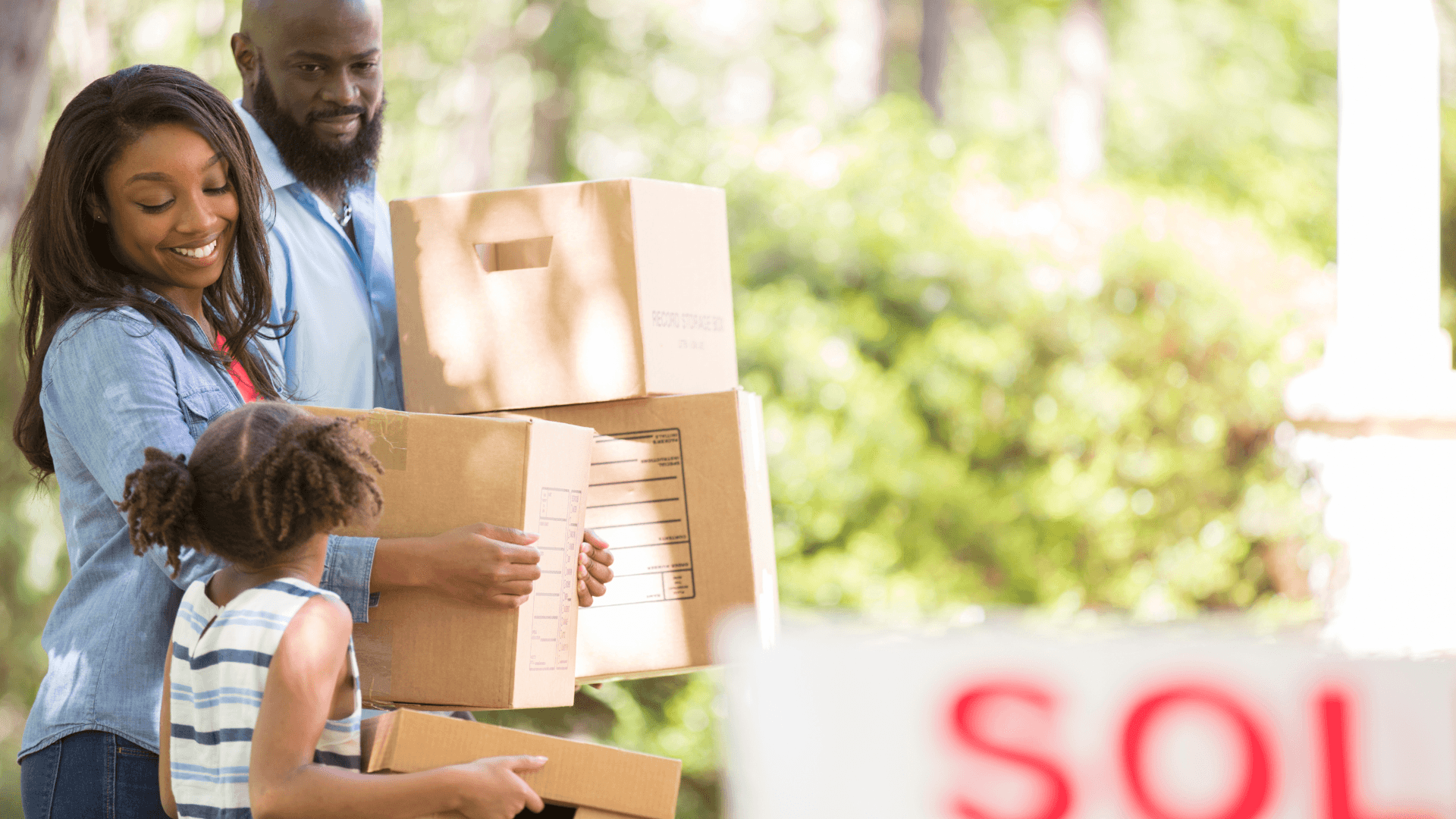 A family carrying boxes past a Sold sign on a summer move
